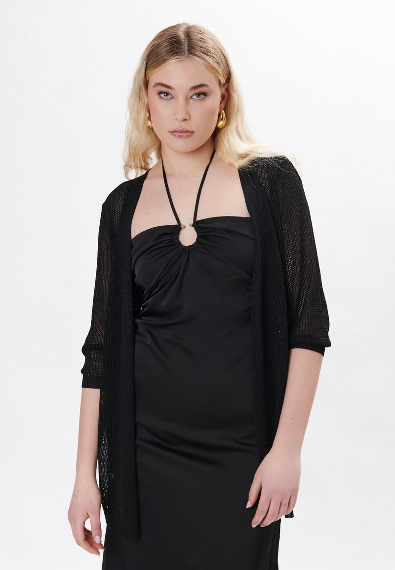 Schwarze, offen gestrickte Strickjacke mit Dreiviertelärmeln über einem schwarzen ärmellosen Kleid, das einen runden Ausschnitt am Kragen aufweist.