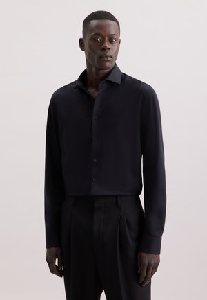 Schwarzes Langarm-Shirt aus glattem Stoff, mit Knopfleiste vorne, klassischem Kragen und tailliertem Schnitt, kombiniert mit passenden Hosen und neutralem Hintergrund.
