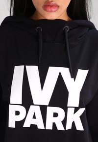Felpa nera con cappuccio, caratterizzata da un grande logo bianco "IVY PARK". Presenta lacci e un tessuto testurizzato, enfatizzando un design casual.
