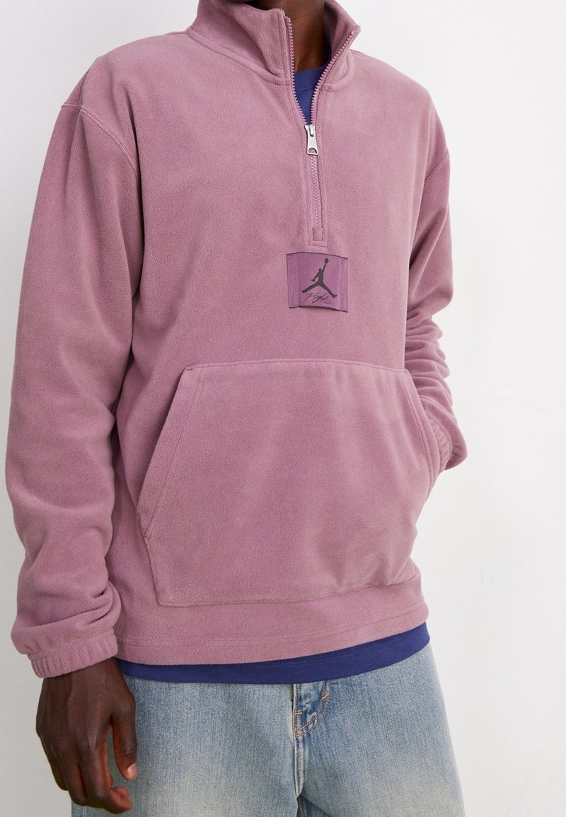 Lila fleece-pull med halv dragkedja, framfickor i kangaroo-stil och en logotypspatch på bröstet. Mjuk textur och avslappnad passform.