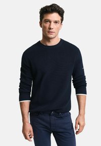 Granatowy sweter z prążkowanego dzianiny z okrągłym dekoltem i kontrastującymi białymi mankietami, noszony z ciemnoniebieskimi spodniami. Teksturowany materiał ukazuje subtelne różnice.