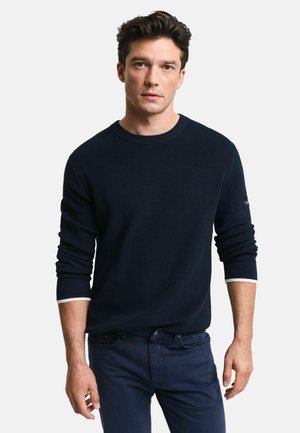 Marineblaues geripptes Strickpullover mit Rundhalsausschnitt und kontrastierenden weißen Bündchen, getragen mit dunkelblauen Hosen. Der strukturierte Stoff zeigt subtile Variationen.