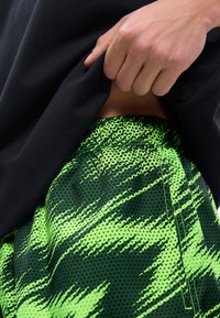 Χέρι που σηκώνει μαύρο πουκάμισο, αποκαλύπτοντας την πράσινη και σκουρόχρωμη ελαστική μέση με σχέδιο σε σορτς ή παντελόνι.