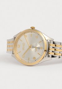 Hopeakeltainen ja kultakeltainen kello minimalistisella suunnittelulla, jossa on sileä kultakeltainen kehys, yksinkertaiset tuntimerkit ja kaksivärinen metalliranneke.