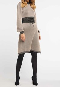 Abito lavorato a maglia beige con maniche lunghe, caratterizzato da una vita nera arricciata e bordo nero ai polsini e all'orlo. Indossato con collant neri e tacchi.