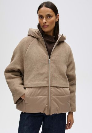 Frau trägt eine beige zweifarbige Reißverschlussjacke mit Wolloberteil und gepolsterten unteren Bereichen, steht mit einer Hand in der Tasche vor einfarbigem Hintergrund.