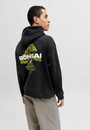 Młody mężczyzna w czarnej bluzie z kapturem z nadrukiem zielonego drzewa bonsai i słowem „BONSAI” na plecach, stojący na tle jasnego, jednolitego tła.
