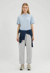 Lichtblauwe t-shirt met een logo, grijze joggingbroek en een marineblauwe sweatshirt die om de taille is geknoopt. Casual sneakers maken de outfit compleet.