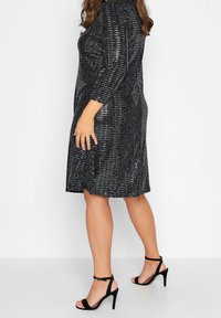 Svart klänning som går till knäna med långa ärmar, med ett texturerat mönster av paljetter; kombineras med svarta högklackade sandaler med remmar.