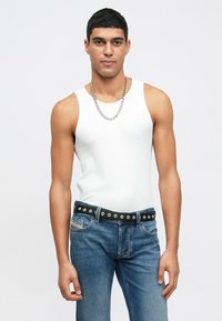 Top de alças brancas sem mangas combinado com jeans de ganga azul. Os jeans têm um cinto preto com studs metálicos e bolsos. O modelo está de frente.