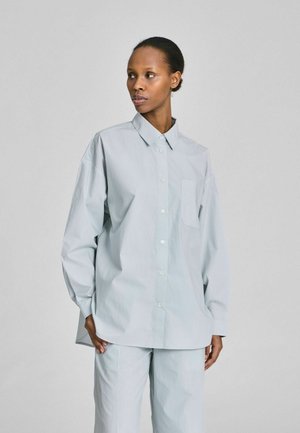 Vrouw draagt een oversized lichtgrijs overhemd met knopen en een borstzak, staand voor een effen lichtgrijze achtergrond.