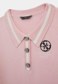 Camisa polo de tejido rosa claro con cuello, tres botones negros y detalles de rayas blancas. Presenta un logotipo bordado en negro.