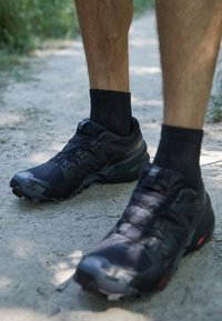Schwarze Sportschuhe mit strukturiertem Synthetik-Obermaterial, robustem Sohlenprofil und Schnürsenkeln, kombiniert mit schwarzen gerippten Socken, auf einem Naturweg.