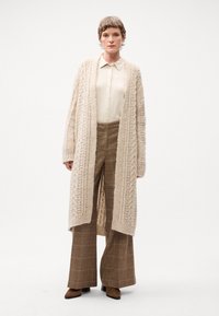 Cardigan lungo beige in maglia con motivo a treccia, indossato su una camicetta crema con bottoni. Pantaloni a quadri marroni a gamba larga e stivali con tacco marroni.