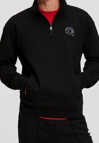 Sudadera negra con media cremallera, de textura suave y con un logo gráfico bordado. Llevada sobre una camiseta roja de cuello redondo, con bolsillos laterales.