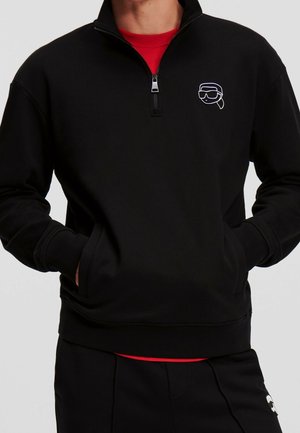 Sudadera negra con media cremallera, de textura suave y con un logo gráfico bordado. Llevada sobre una camiseta roja de cuello redondo, con bolsillos laterales.