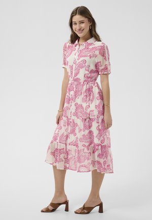 Młoda kobieta uśmiechająca się, ubrana w białą sukienkę midi z różowym wzorem paisley, krótkimi rękawami, z przodem na guziki oraz brązowymi sandałami na obcasie.