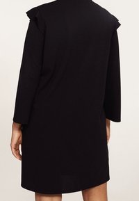 Vestido negro con mangas tres cuartos, con acentos de hombros con volantes. Textura simple y suave, diseño a la altura de la rodilla y corte relajado.