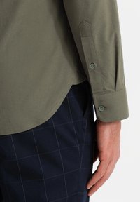 Olijfgroene shirt met knoopmanchetten, met een gladde textuur en een afgeronde zoom. Gecombineerd met marineblauwe geruite broek, zichtbaar aan de onderkant.