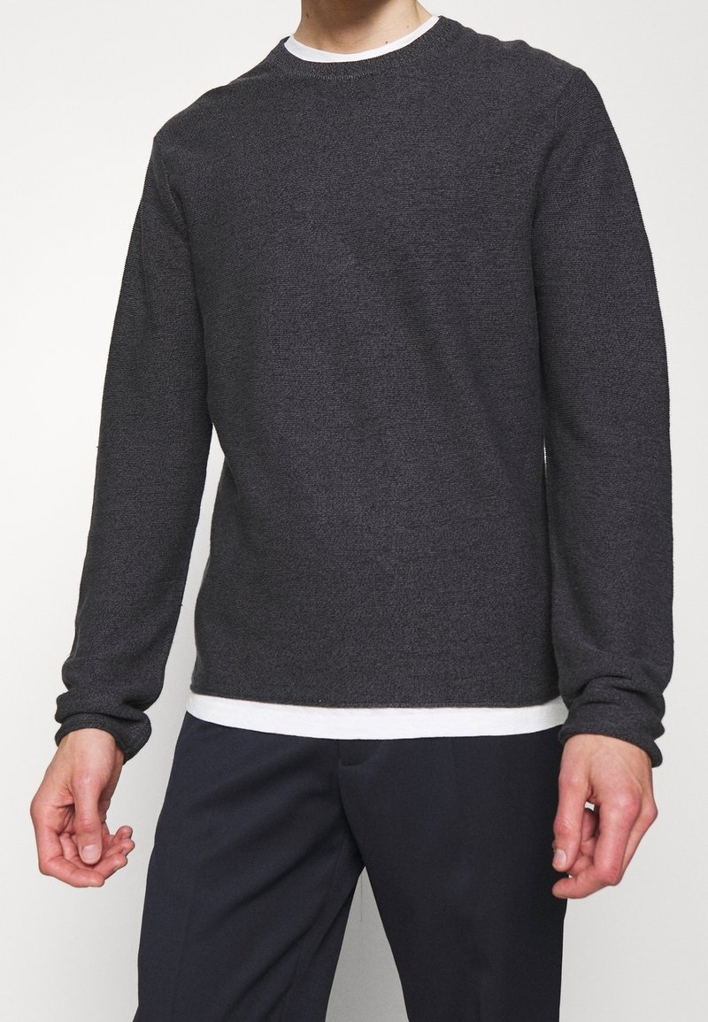 Homme portant un pull à manches longues gris foncé par-dessus une chemise blanche avec un pantalon noir sur un fond blanc uni.