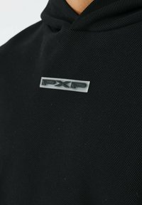 Sudadera negra con una tela texturizada, que presenta un pequeño logo "PXP" en plata y negro en el frente. Tiene un cuello doblado y un diseño aerodinámico.