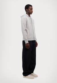 Lysegrå hettejakke med glidelås, snorhette, sidelommer og ribbestrikkede mansjetter, kombinert med mørkeblå løse bukser og beige sneakers.