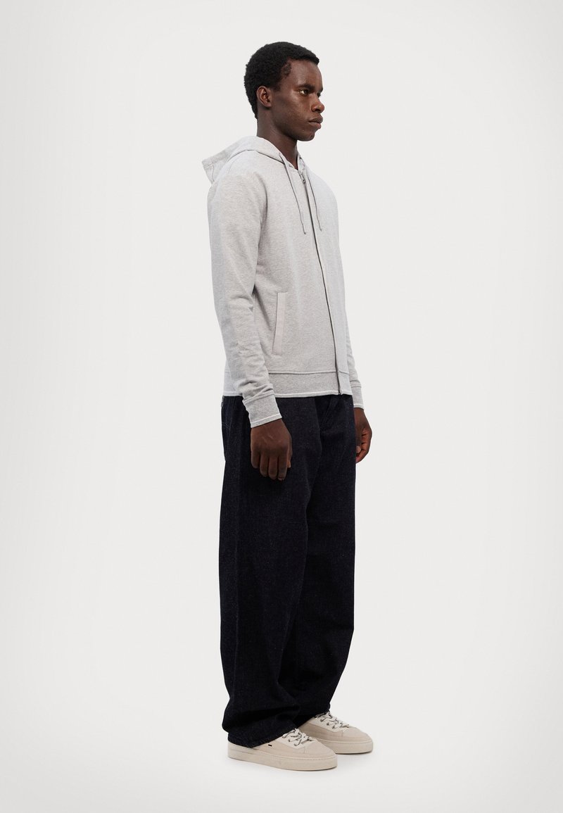 Lysegrå hettejakke med glidelås, snorhette, sidelommer og ribbestrikkede mansjetter, kombinert med mørkeblå løse bukser og beige sneakers.