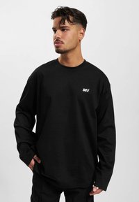 Sort langærmet T-shirt lavet af bomuld, med rund halsudskæring og et lille hvidt "DEF" logo på den venstre brystområde.