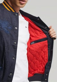 Veste bomber en denim avec broderie de dragon bleu, doublure matelassée rouge, col rehaussé de jaune et fermeture à boutons-pression.