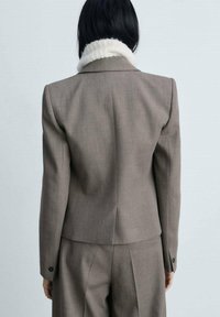 Räätälöity ruskea bleiseri, jossa on valkoinen tekoturkiskaulus, muodollinen istuvuus, pitkät hihat ja napitettavat hihansuut, valmistettu sileästä kankaasta.