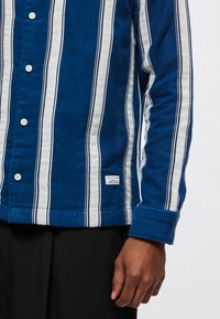 Niebiesko-biała koszula na guziki w paski, wykonana z miękkiego materiału, z kołnierzykiem i metką z boku na dole, zestawiona z czarnymi spodniami.