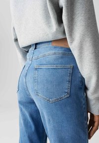 Persona che indossa jeans in denim azzurro chiaro e un felpa grigia, mostrata di spalle con una mano rilassata lungo il fianco.