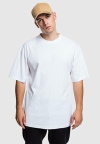 Witte oversized katoenen t-shirt met korte mouwen, ronde hals; gecombineerd met een tan baseballcap, donkere broek, tegen een grijze achtergrond.