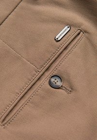 Tissu texturé couleur fauve avec une poche à boutons et une petite étiquette métallique marquée « QUINTESSENCE » près de la couture de la poche.