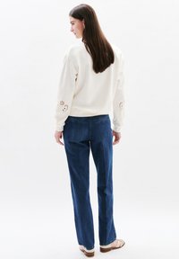 Felpa bianca con dettagli ricamati sulle maniche, abbinata a jeans blue e sandali chiari, vista da dietro su uno sfondo neutro.