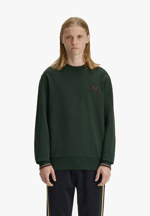 Persona giovane con capelli alla lunghezza delle spalle che indossa una felpa verde scuro e pantaloni neri con strisce laterali gialle, in piedi contro uno sfondo semplice.