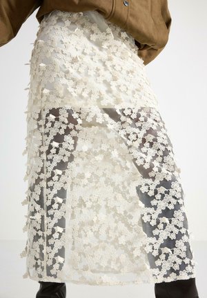 Persona che indossa una gonna di pizzo bianco trasparente con motivi floreali sopra collant neri e una giacca marrone, in piedi davanti a uno sfondo semplice.