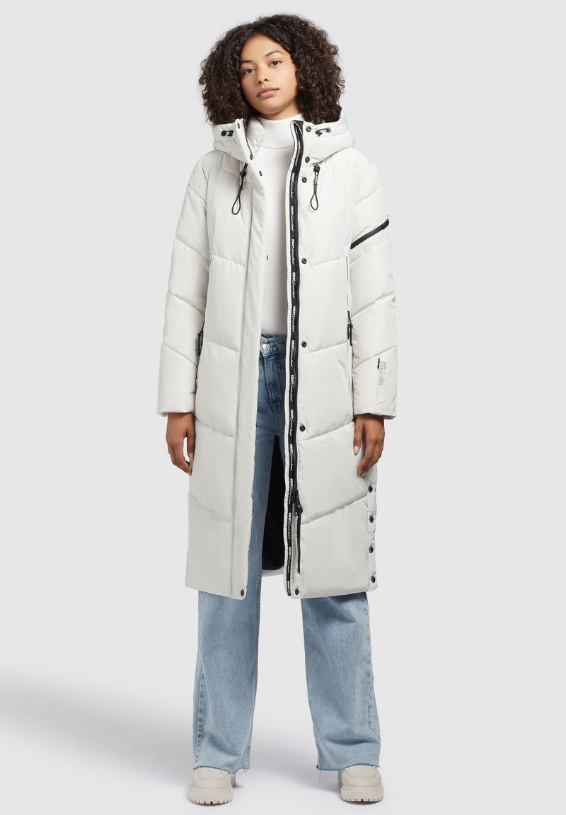 Weißer Puffer-Mantel mit Kapuze, mit einem Reißverschluss vorne, Druckknopfverschluss und diagonalen schwarzen Streifen an den Ärmeln. Trägt er über heller Jeans.