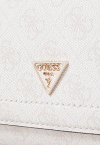 Hvid tekstureret håndtaske med en guld trekantet logo-plade mærket "GUESS USA" med et spørgsmålstegn og synlige sy-detaljer.