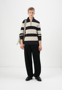 Gestreifter beige und marineblauer Pullover mit einem Viertelreißverschluss, kombiniert mit schwarzen, locker sitzenden Hosen und schwarzen Schuhen, getragen mit einer grauen Schultertasche.
