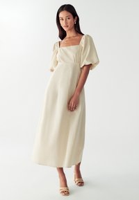 Bežs puff-sleeve midi kleita ar kvadrātveida dekoltē, pieguļošu augšdaļu un teksturētu audumu. Valkāta ar plikām papēžkurpēm.