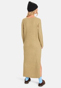 Beiges, geripptes Strickkleid mit langen Ärmeln, Seitenschlitzen und lockerer Passform. Getragen mit einer schwarzen Mütze und braunen Schuhen über blauen Socken.