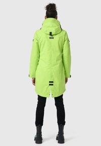 Groene waterdichte parka met capuchon, voorzien van zwarte hardware en accenten, een lange snit en verstelbare elementen, in combinatie met zwarte broek en laarzen.