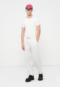 Camiseta blanca, pantalones de chándal grises con cintura elástica y zapatos negros de suela gruesa. Modelo lleva una gorra de béisbol roja y negra.