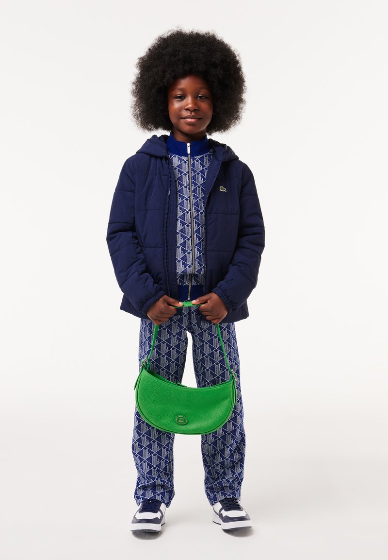 Doudoune bleu marine, survêtement à motifs bleus, sac à main texturé vert et baskets blanches. Enfant se tient debout avec le sac sur l'épaule.