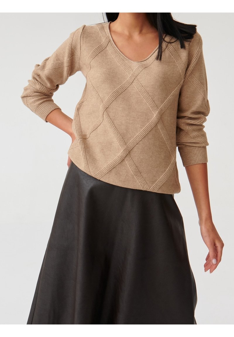 Femme portant un pull beige en V avec un motif torsadé diagonal et une jupe noire en forme A, posant avec une main sur la hanche.