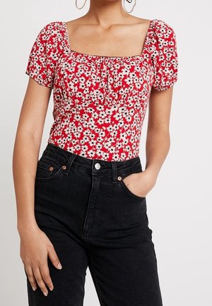 Kobieta ubrana w czerwoną kwiecistą bluzkę z krótkim rękawem i kwadratowym dekoltem oraz czarne jeansy z wysokim stanem, jedna ręka w kieszeni.