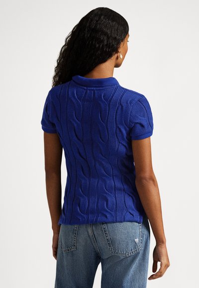 Modra pletena polo majica z teksturiranim vzorcem kablovja. Kratki rokavi in ovratni izrez v kombinaciji s svetlo modrimi jeans hlačami.