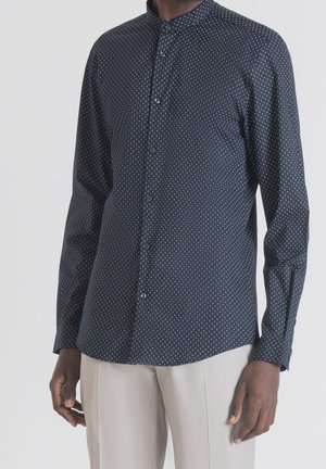 Homme portant une chemise bleu foncé à manches longues avec de petits points blancs et un pantalon beige clair, debout les mains détendues le long du corps.