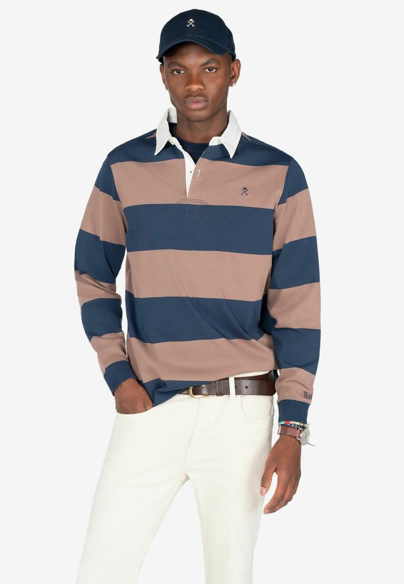 Polo a maniche lunghe a righe blu navy e beige con colletto bianco, dotato di un piccolo logo sul petto e di un tessuto strutturato. Indossato con pantaloni chiari.